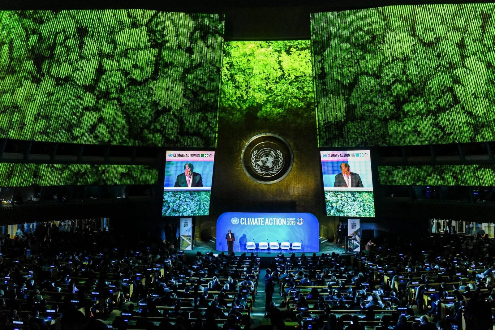Delegates fill the UN General Assembly hall at a climate summit, forest imagery projected above the stage.