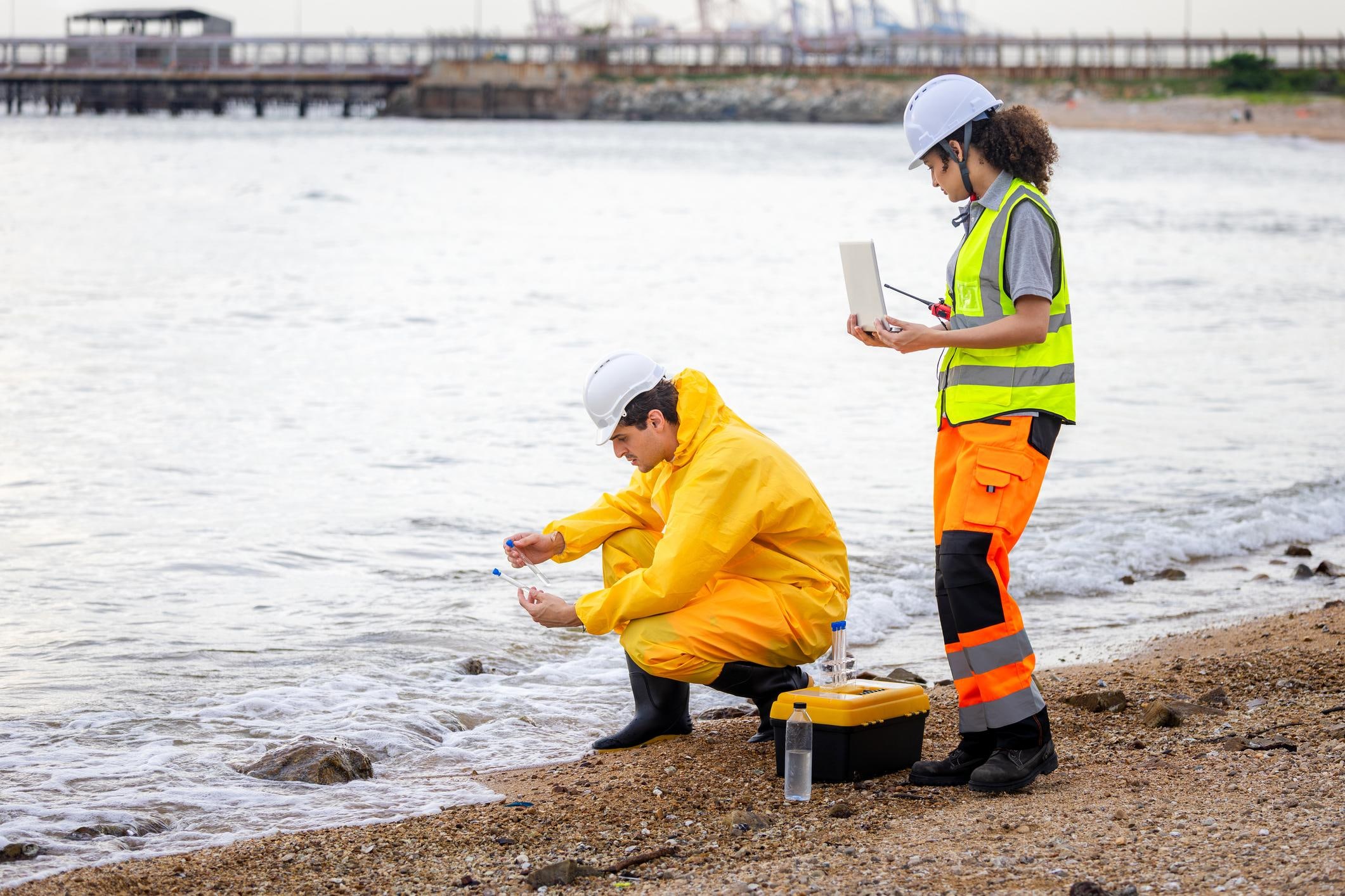 Two technicians in high-visibility gear taking water samples at a shoreline