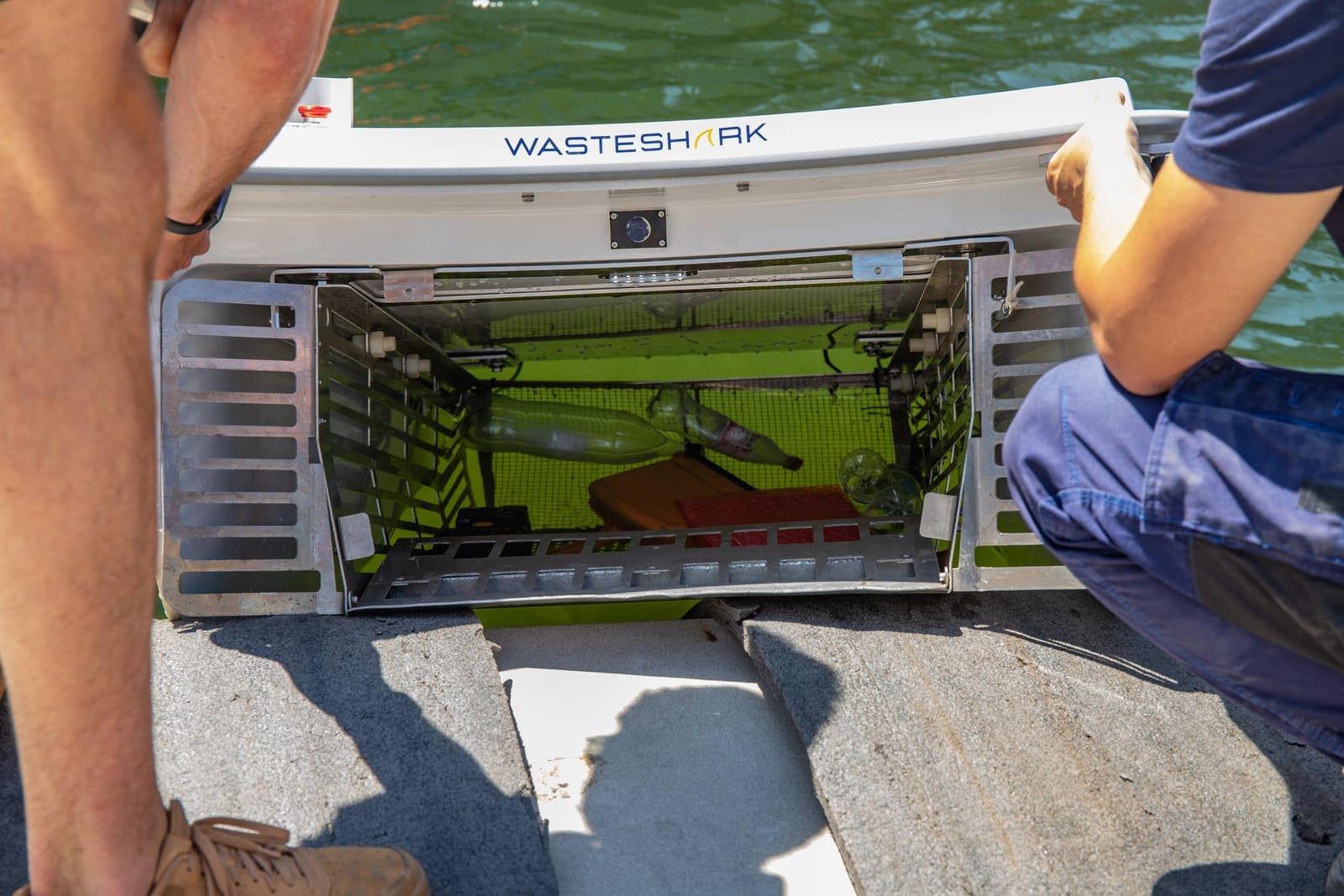 An open WasteShark collection basket filled with plastic bottles and litter, two people looking in.