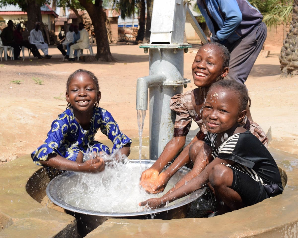 Three children laughing as they splash water from a village handpump into a metal basin